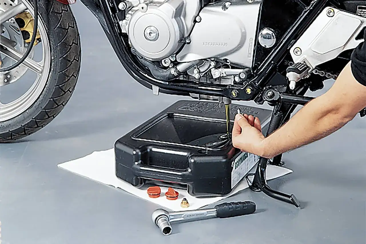 Mano che svita il tappo per cambiare l'olio di lubrificazione del motore di una moto, con una bacinella di raccolta sotto.