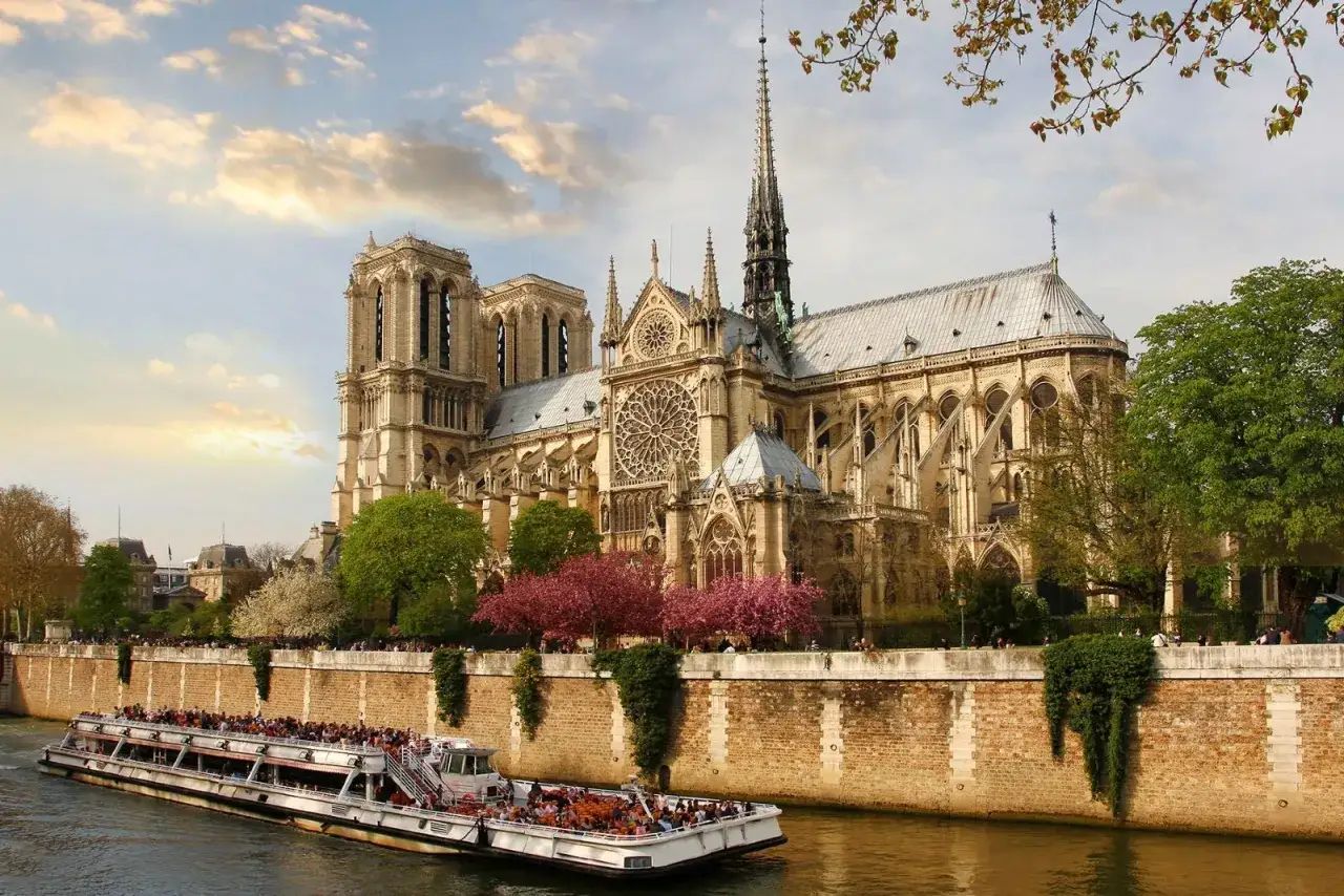 Barco tur&iacute;stico navega por el Sena, con la majestuosa Notre Dame y sus entradas de fondo, rodeada de &aacute;rboles en flor.