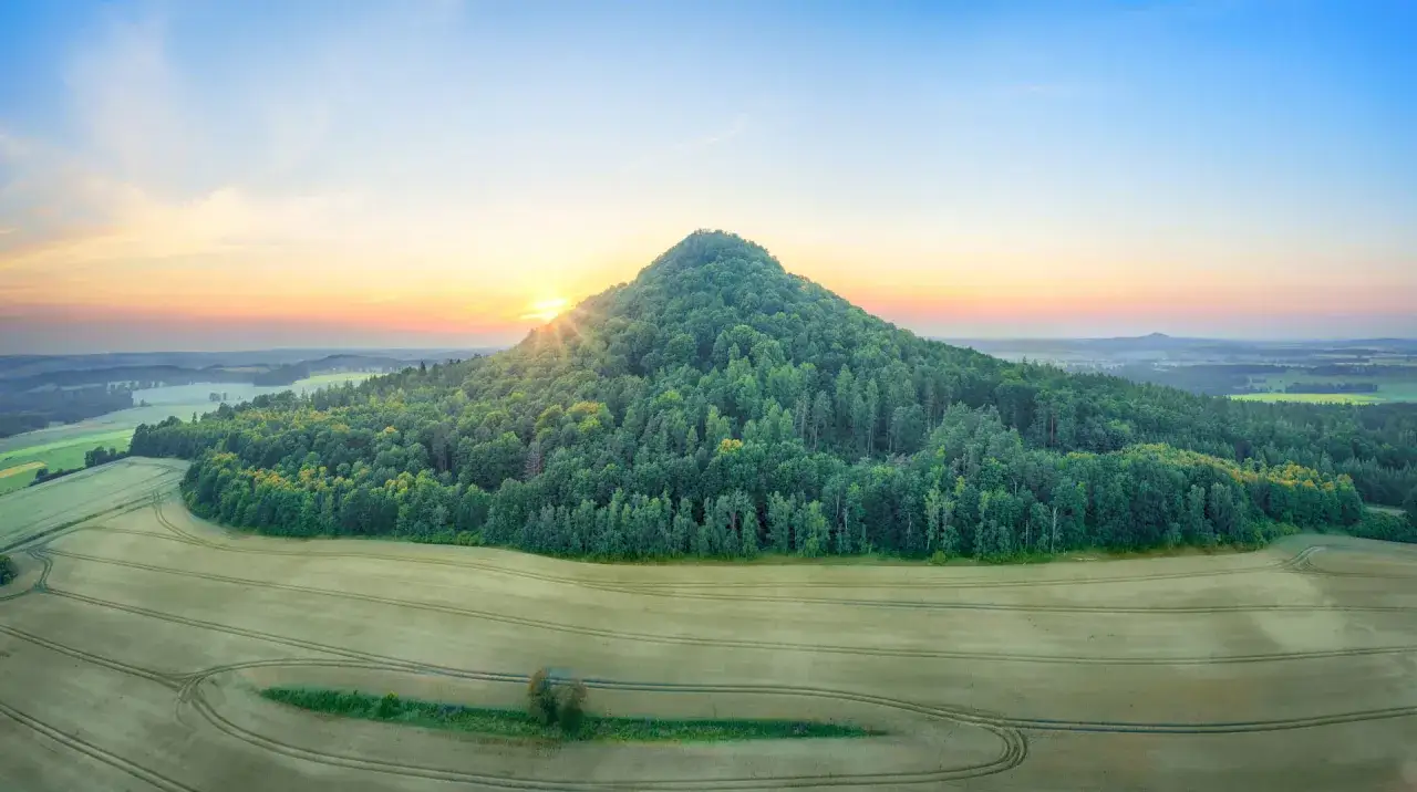 Wzgórze pokryte lasem, dawne ognisko wulkaniczne, stanowi główną atrakcję krajobrazu. Wschód słońca maluje niebo nad polami.