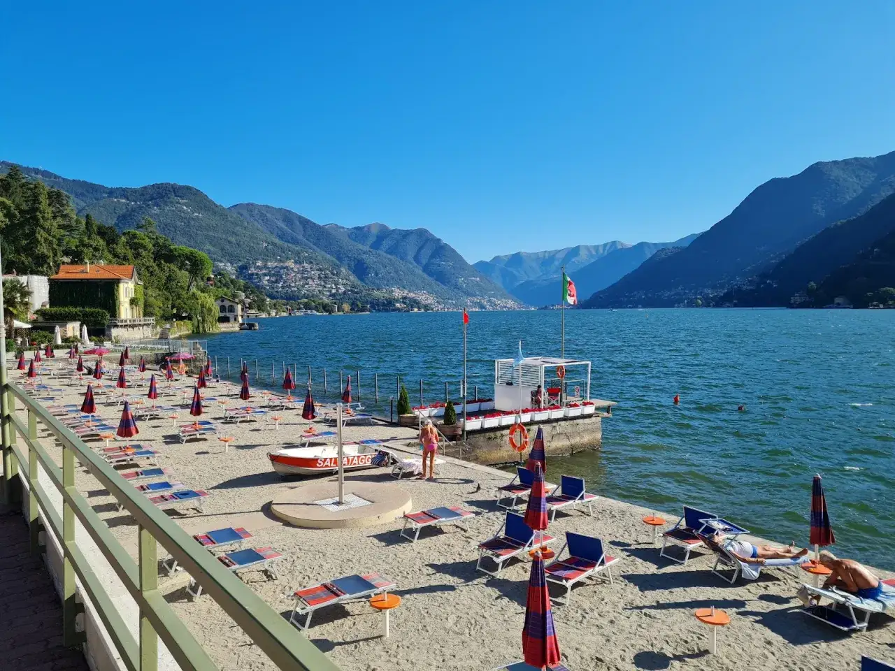 Słoneczne plaże nad jeziorem Como, z rzędami leżaków i parasoli, zapraszają do relaksu. W tle góry i błękitne niebo.