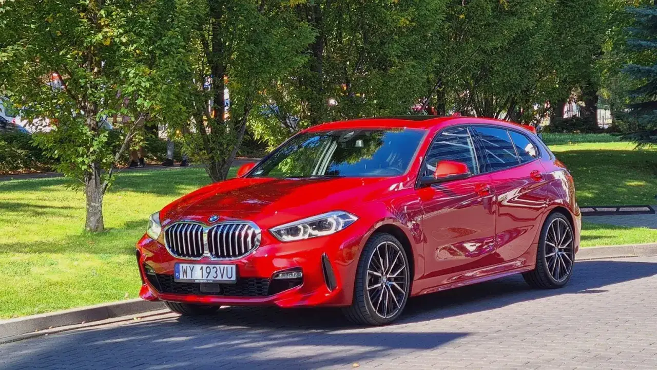 Błyszczące czerwone BMW 118i zaparkowane na tle zieleni i drzew.
