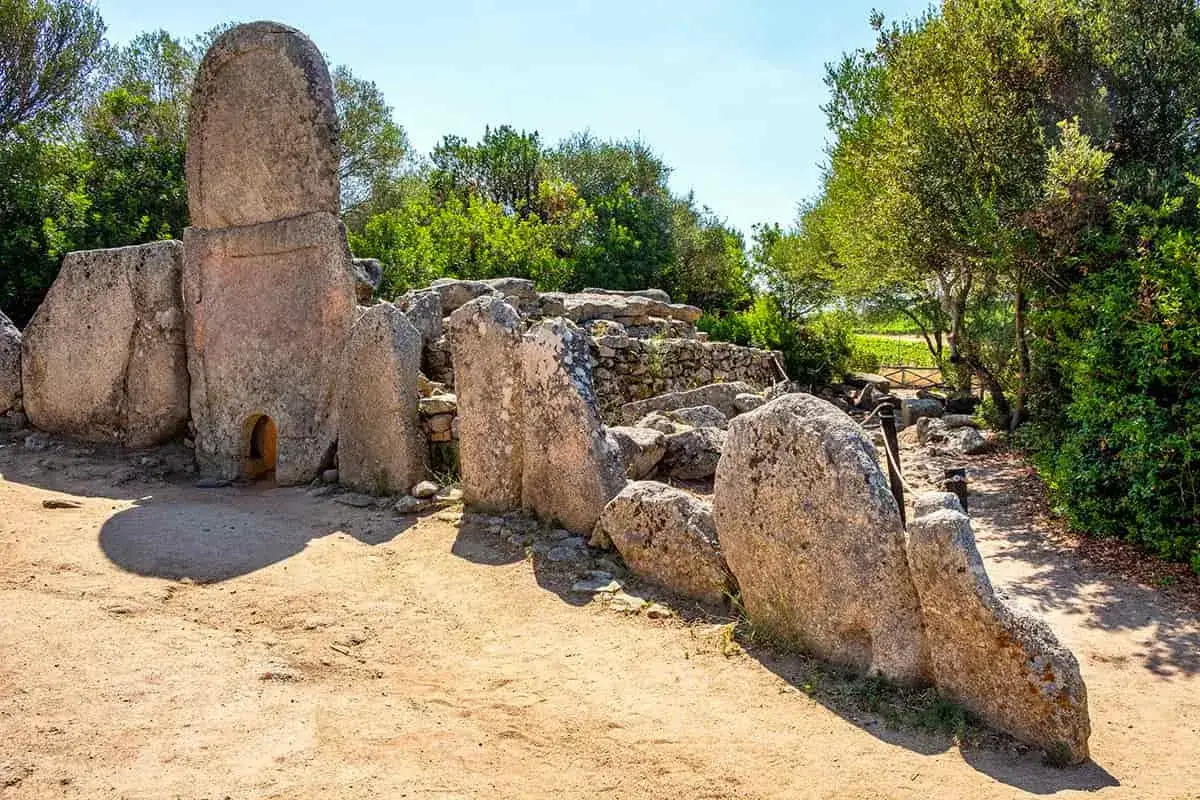 Antiche tombe dei giganti in pietra, con un ingresso ad arco, circondate da vegetazione e un sentiero sabbioso.