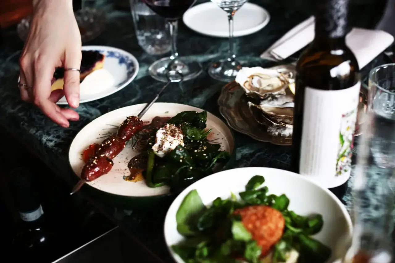 En hand str&auml;cker sig efter en spettad k&ouml;ttbit p&aring; en tallrik med gr&ouml;nsaker. Bredvid finns ostron, vin och en sallad. En h&auml;rlig m&aring;ltid, kanske p&aring; Odenplan.