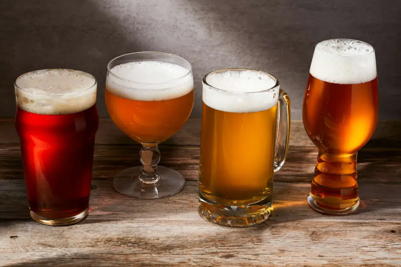 Quatre verres de bière aux couleurs variées, du rouge ambré au doré clair, sur une table en bois rustique.