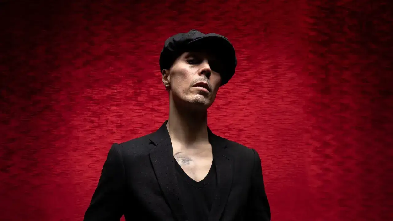 Ville Valo w czarnym garniturze i kaszkiecie na tle czerwonego aksamitu.