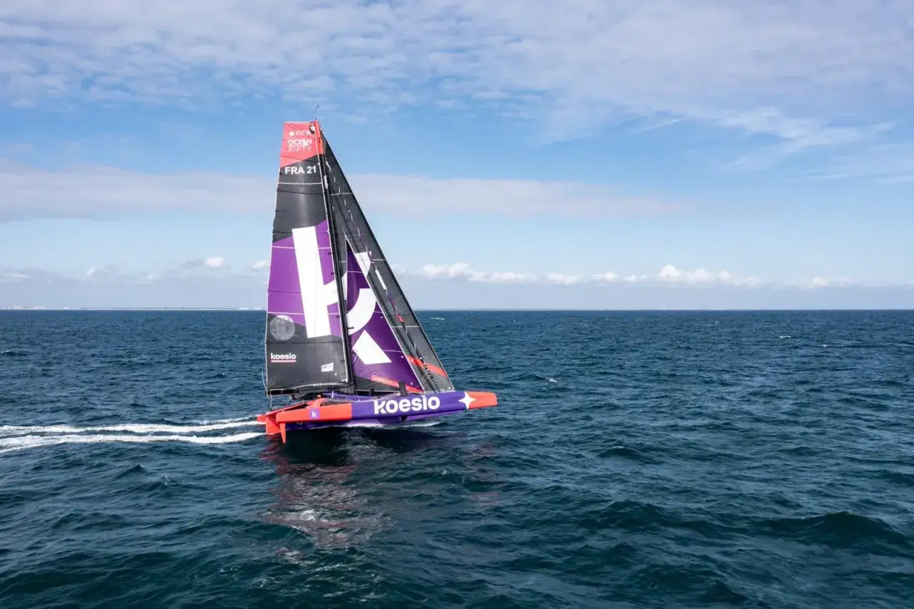 Le trimaran Koesio fend les vagues bleues sous un ciel clair, ses voiles violettes et noires portant le logo de la course Ocean Fifty.