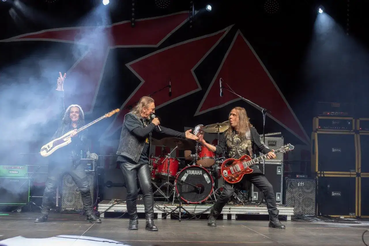 Rockowi giganci, TSA dream team, na scenie. Wokalista śpiewa, gitarzyści grają, perkusista w tle.