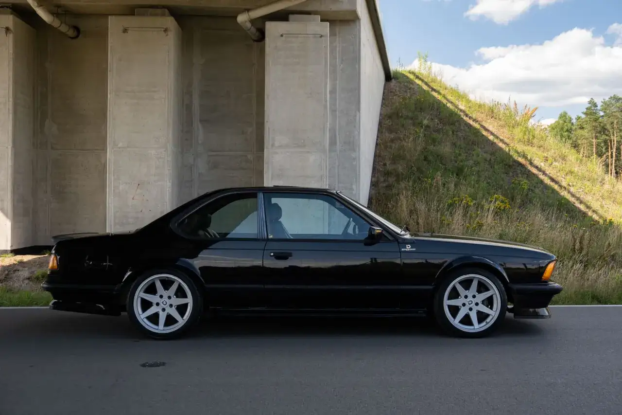 Czarny, lśniący **bmw 635 csi** z lat 80. stoi pod betonowym wiaduktem, obok porośniętego trawą nasypu.