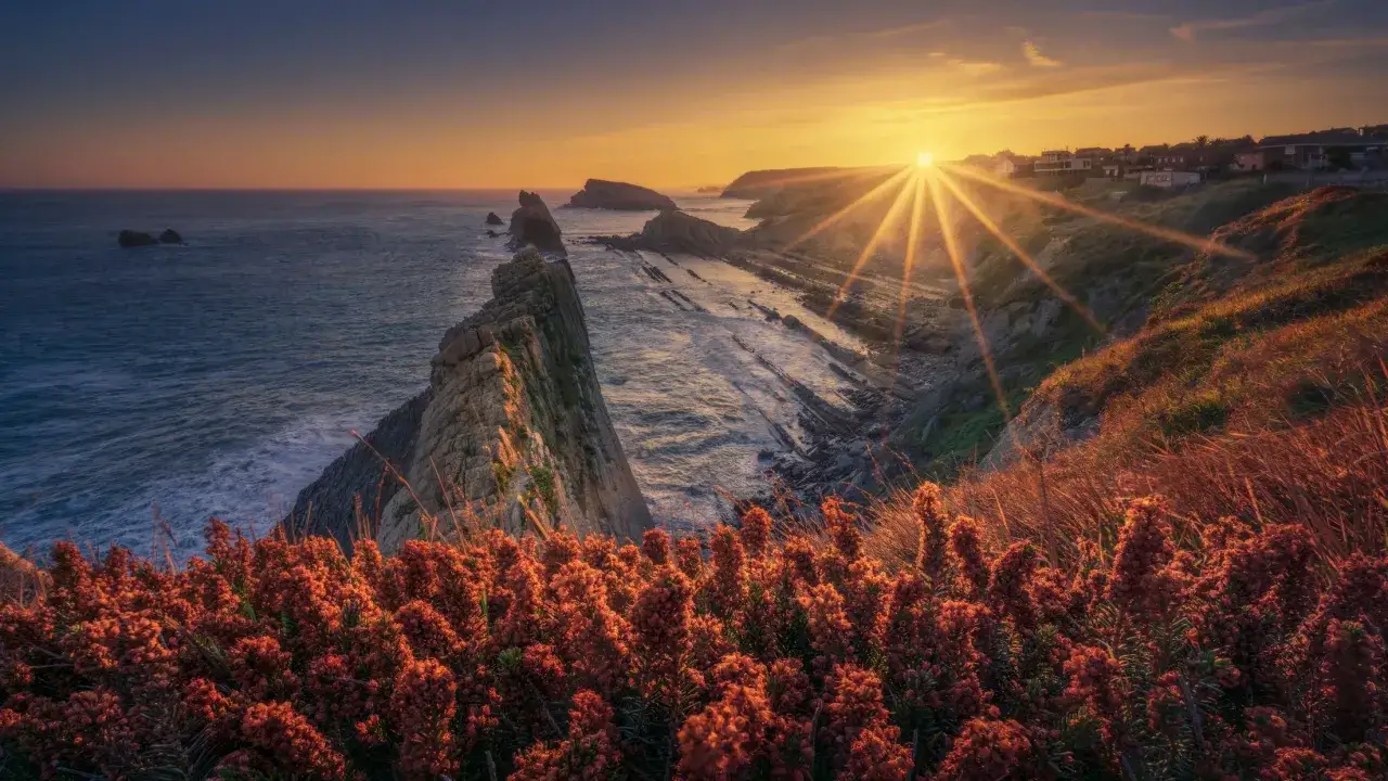 Atardecer dorado sobre los acantilados de Urros de Liencres. El sol se pone entre las rocas y el mar, iluminando la costa y la vegetaci&oacute;n.