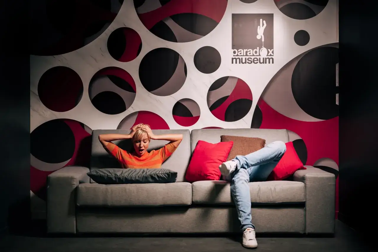 Une femme choquée sur un canapé devant un mur de cercles. Le logo du Paradox Museum est visible. La durée de visite du musée de l'illusion est une expérience inoubliable.