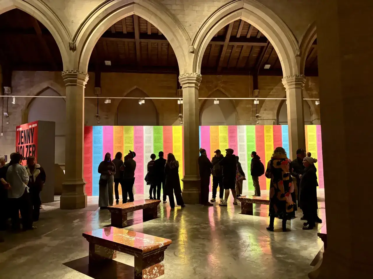 Visitors admire a vibrant wall of textual art, a colorful display of Jenny Holzer's work, in a grand arched hall.