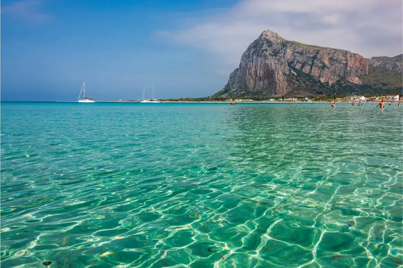 Acque cristalline e turchesi del mare di San Vito Lo Capo, con la maestosa montagna a fare da sfondo e barche a vela all'orizzonte.