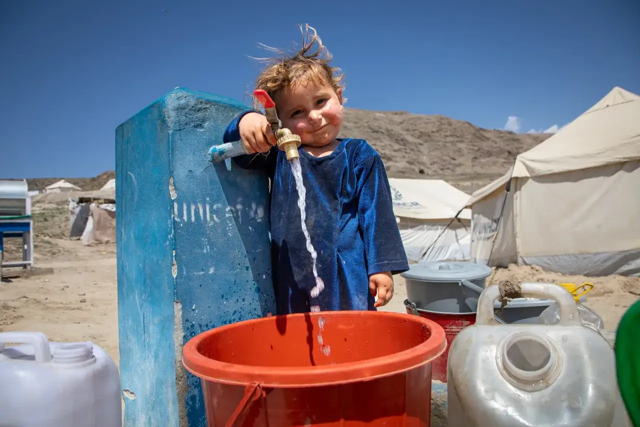 Ett litet barn i blå klänning samlar rena vatten från en kran vid en UNICEF-station. Tält och berg syns i bakgrunden.