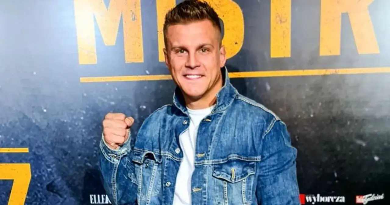 Uśmiechnięty Alan Kwieciński w dżinsowej kurtce pozuje z zaciśniętą pięścią na tle napisu.