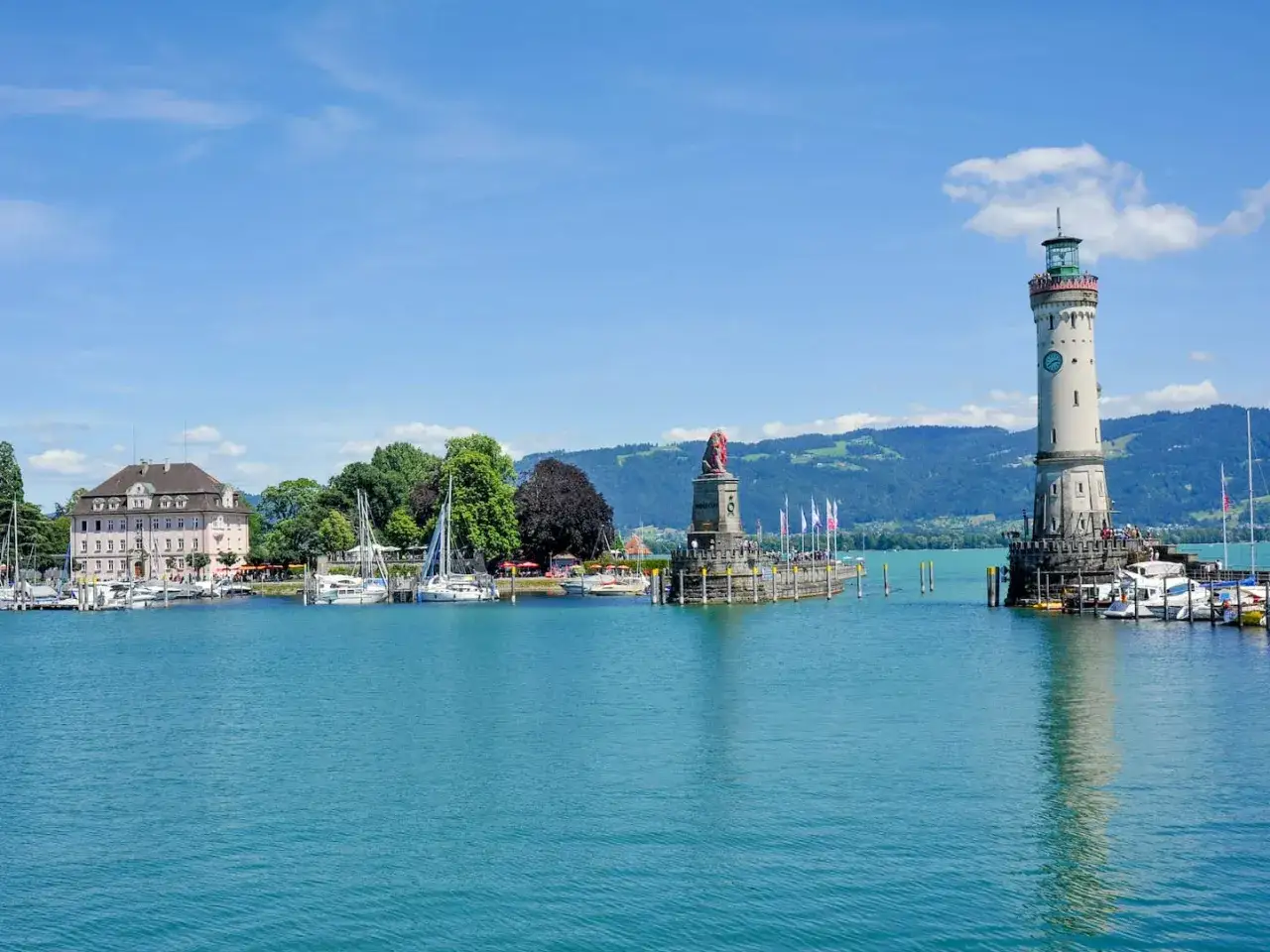 Der Leuchtturm Lindau wacht &uuml;ber den Hafen mit dem L&ouml;wendenkmal. Boote liegen im t&uuml;rkisfarbenen Wasser vor Anker.