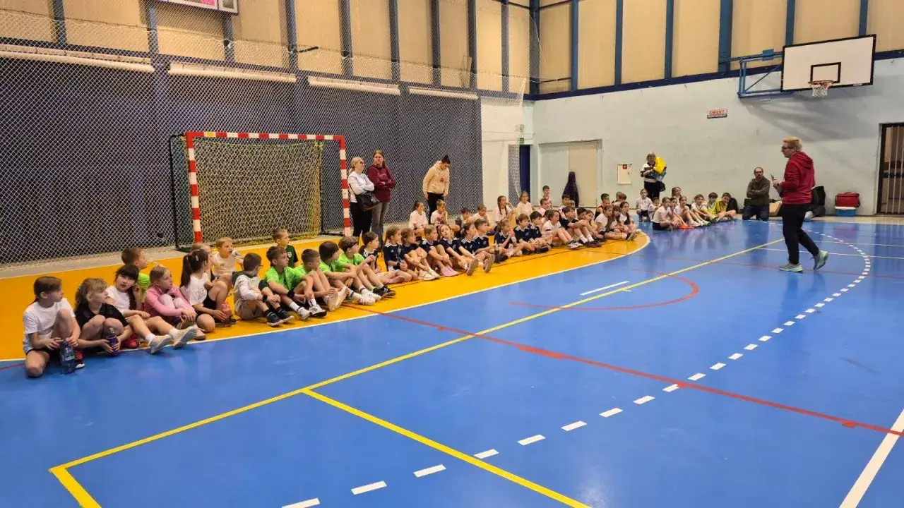 Dzieci z MUKS Zagłębie Sosnowiec siedzą na hali sportowej, słuchając trenera.