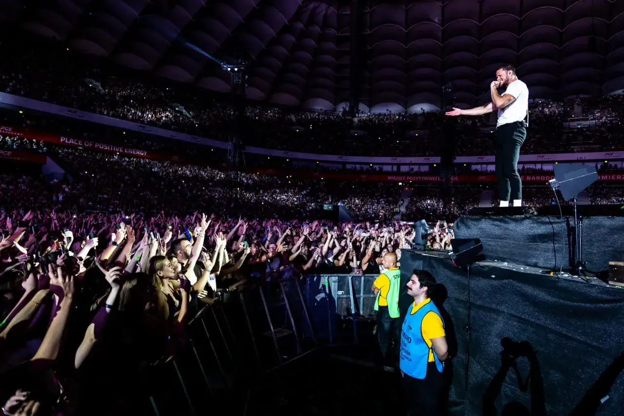 Koncert Imagine Dragons na PGE Narodowym. Wokalista śpiewa do tłumu, który szaleje z telefonami w górze.