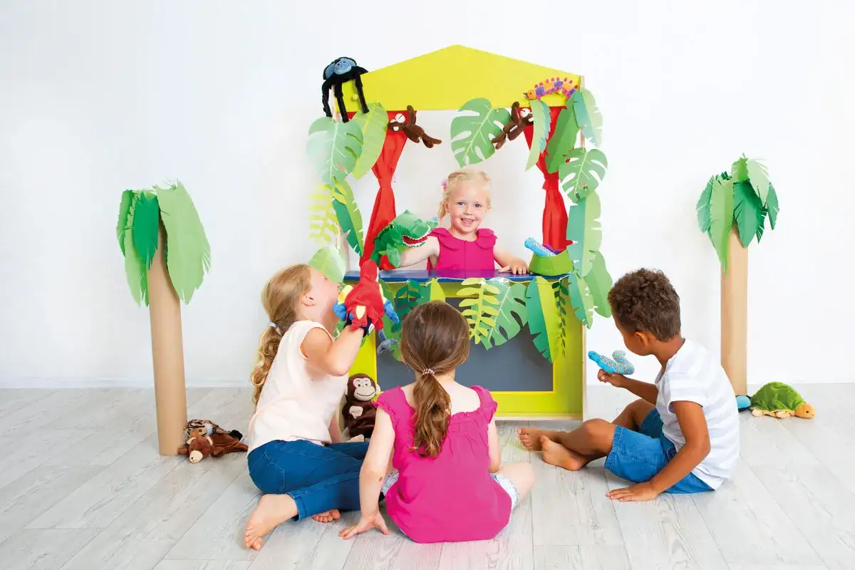 Kinder spielen im **Vorlesetheater Grundschule** mit Handpuppen im Dschungel-Design. Ein M&auml;dchen im gelben Theaterst&uuml;ck h&auml;lt einen gr&uuml;nen Dinosaurier.