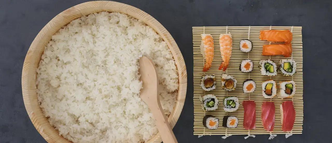 Gotowy ryż do sushi w drewnianej kadzi, obok mata bambusowa z r&oacute;żnymi rodzajami sushi. Zastanawiasz się, ile octu do sushi?
