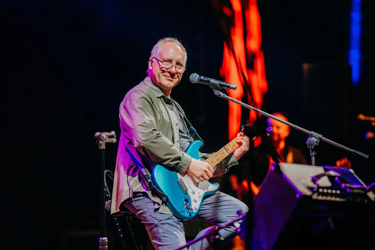 Mężczyzna gra na niebieskiej gitarze elektrycznej na scenie. Koniec tekst.
