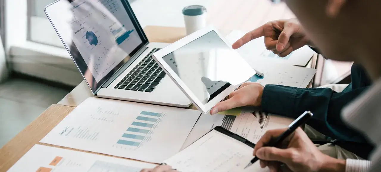 Deux personnes analysent des graphiques sur un ordinateur portable et une tablette, signe d'une gestion administrative d'entreprise efficace.
