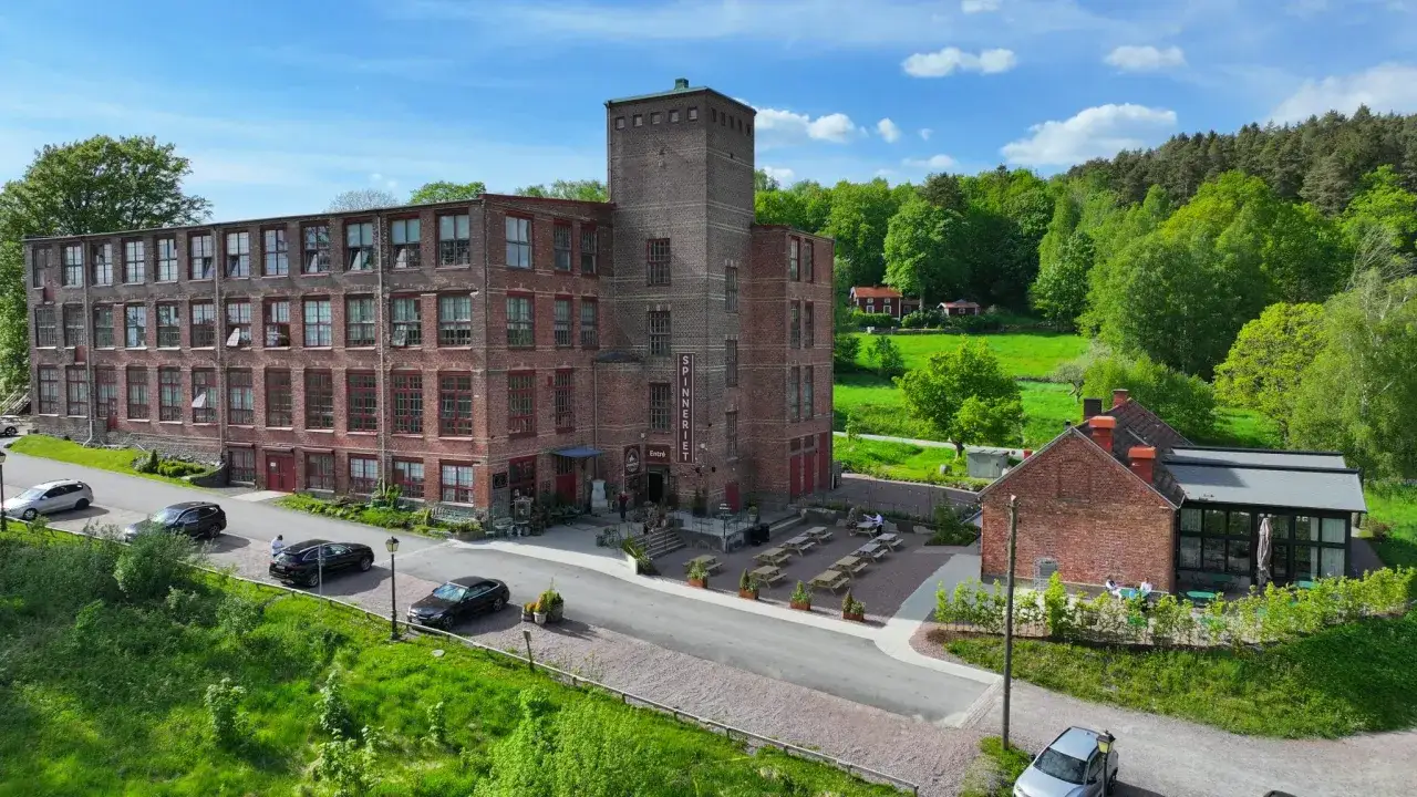 Lindome Spinneri, en historisk industribyggnad i tegel, omgiven av grönskande natur. Utomhusbord och stolar inbjuder till avkoppling.