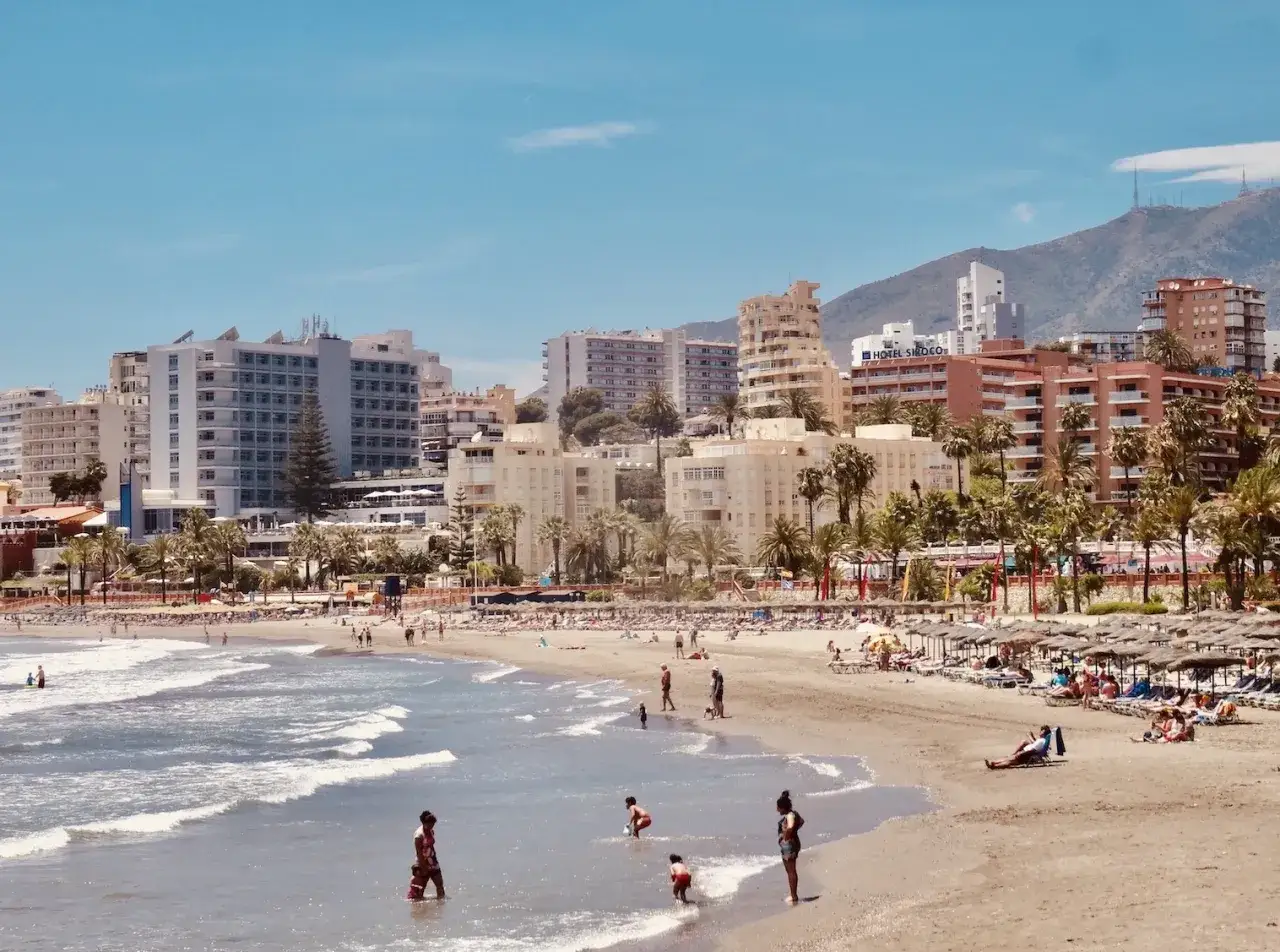 Sonniger Benalmadena Strand mit vielen Menschen, Hotels und Palmen. Kinder planschen im Meer, w&auml;hrend Urlauber auf Liegest&uuml;hlen entspannen.
