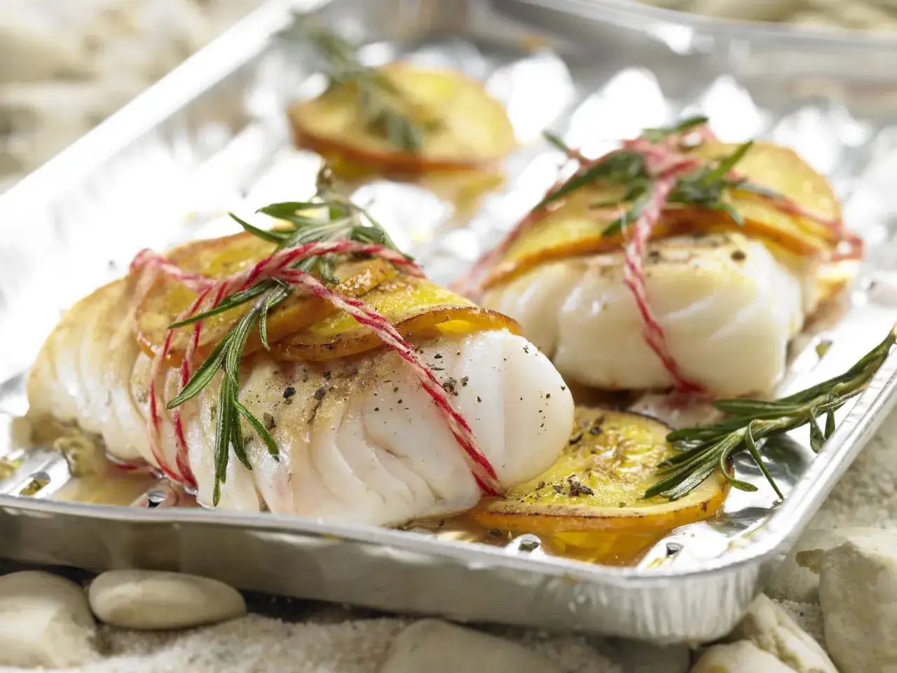 Zwei Filets Kabeljau, mit Zitronenscheiben und Rosmarin umwickelt, werden auf einem Blech gegrillt. Ein köstliches Gericht, kabeljau grillen ohne alufolie.