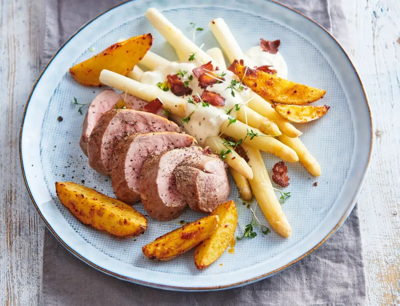 Saftiges schweinefilet mit spargel, Kartoffelspalten und einer cremigen Sauce, garniert mit Speck und Kr&auml;utern.