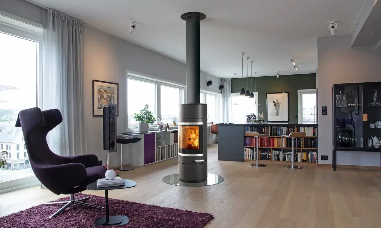 Modern vardagsrum med en centralt placerad **inbyggnadskamin** som sprider v&auml;rme. En m&ouml;rklila f&aring;t&ouml;lj och en bokhylla full av b&ouml;cker bidrar till mysfaktorn.