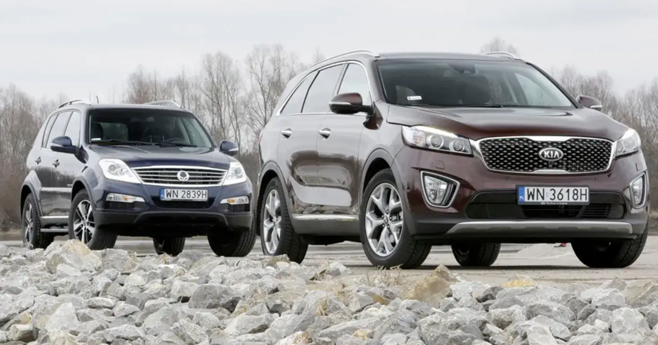 Dwa SUV-y: ciemnoniebieski SsangYong Rexton i bordowy Kia Sorento, stoją na tle kamieni i drzew.