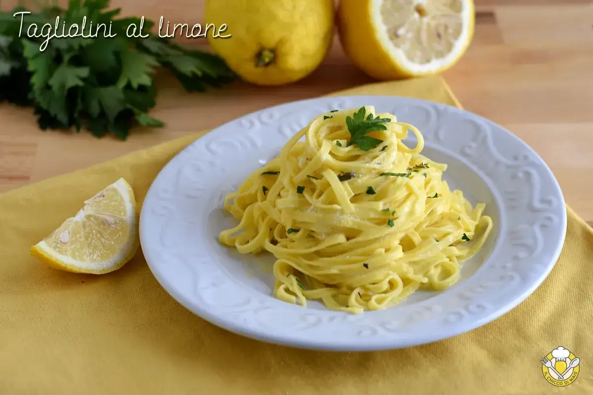 Un piatto di tagliolini al limone, conditi con una salsa cremosa e guarniti con prezzemolo fresco. Accanto, spicchi di limone e foglie di prezzemolo.