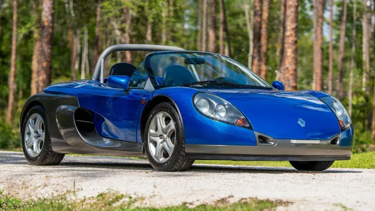 Niebieski Renault Wind, sportowy kabriolet, zaparkowany na żwirowej drodze wśród drzew.