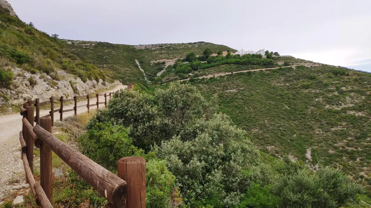 Sendero de monta&ntilde;a con vallas de madera, vegetaci&oacute;n exuberante y edificios blancos en la cima. Un tramo del camino de Santiago Pe&ntilde;&iacute;scola.