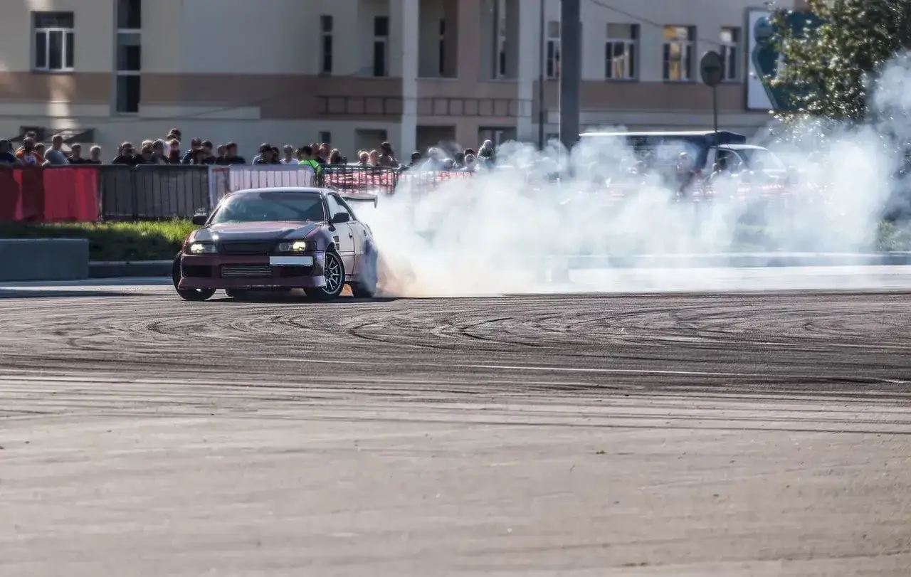 Samochód sportowy w kłębach dymu podczas widowiskowego driftu. Widzowie obserwują zmagania, ale kierowca pewnie zmierza po swój mandat za driftowanie.