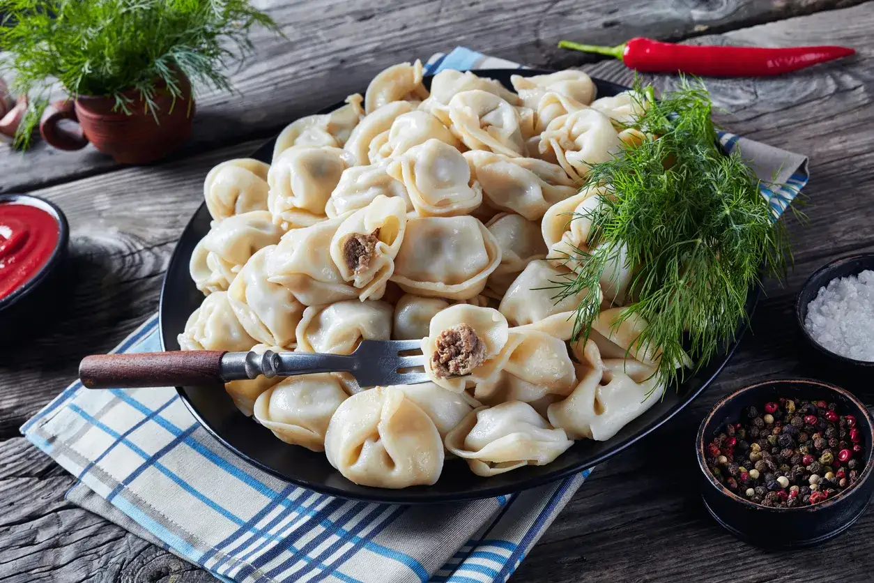 Talerz pełen parujących uszek z mięsem, udekorowany koperkiem. Obok miseczka z sosem, przyprawy i papryczka chili.