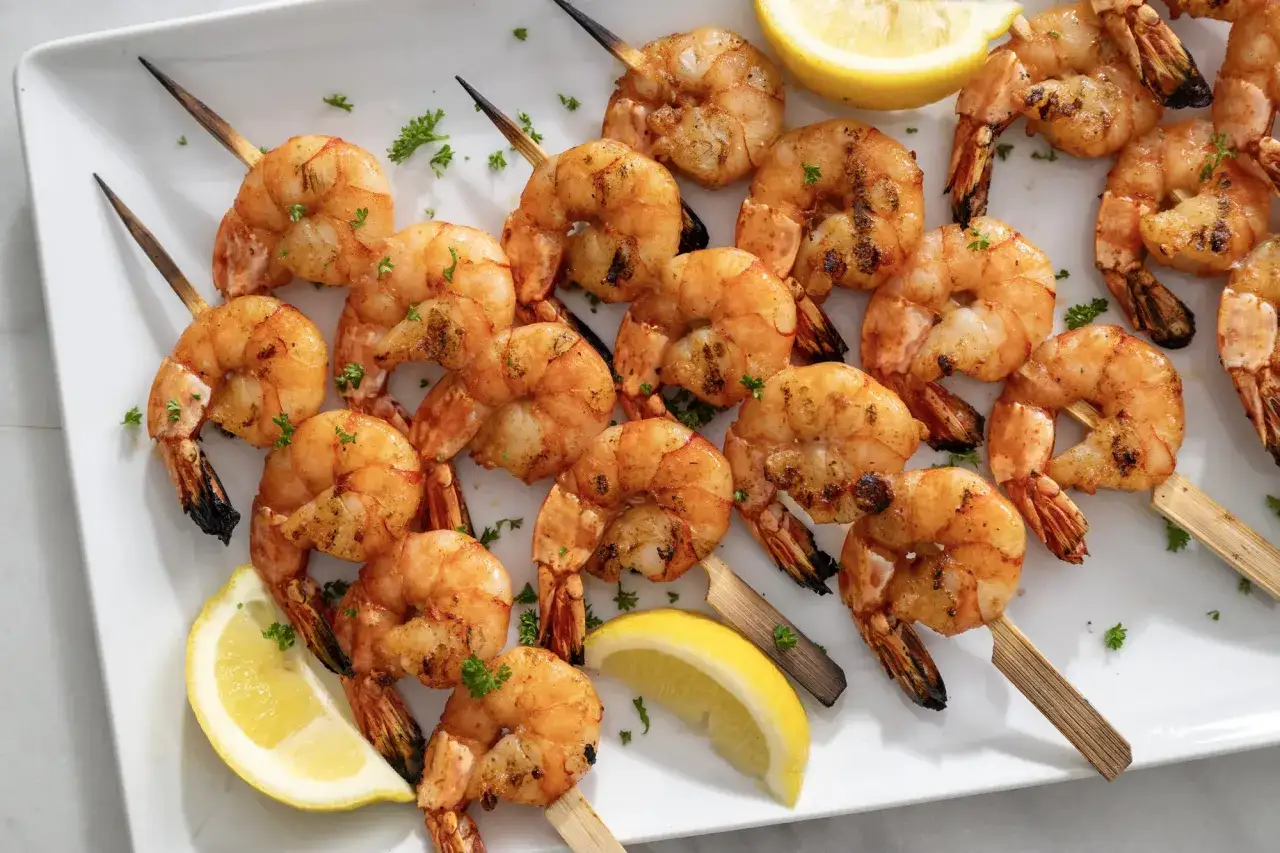 A platter of perfectly grilled shrimp on skewers, garnished with fresh parsley and lemon wedges.
