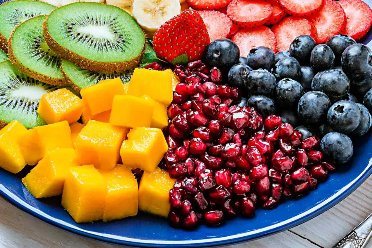 En bl&aring; tallrik fylld med f&auml;rgglad c vitamin frukt: skivad kiwi, mangot&auml;rningar, granat&auml;ppelk&auml;rnor, bl&aring;b&auml;r, jordgubbar och banan.