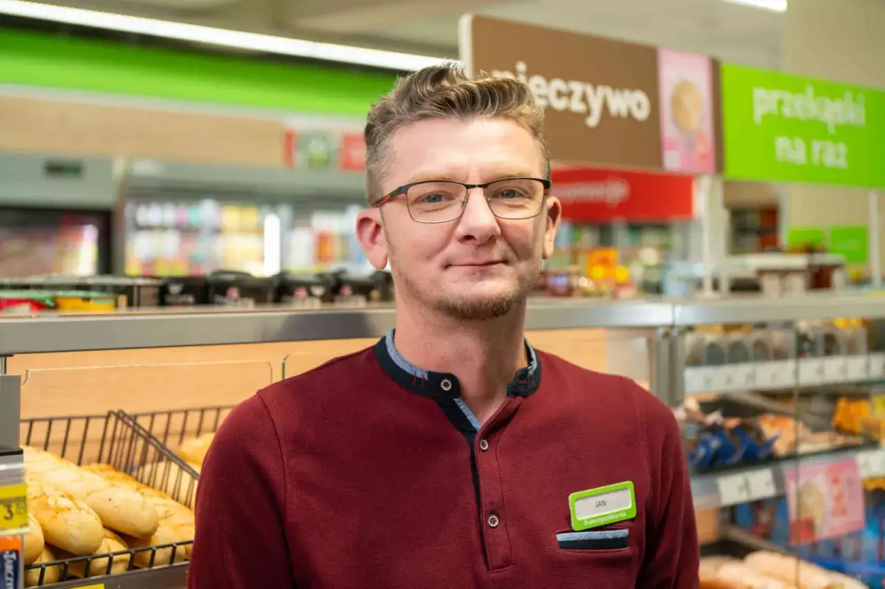 Jan, kierownik Żabki, uśmiecha się do kamery. W tle widać półki z pieczywem i przekąskami.