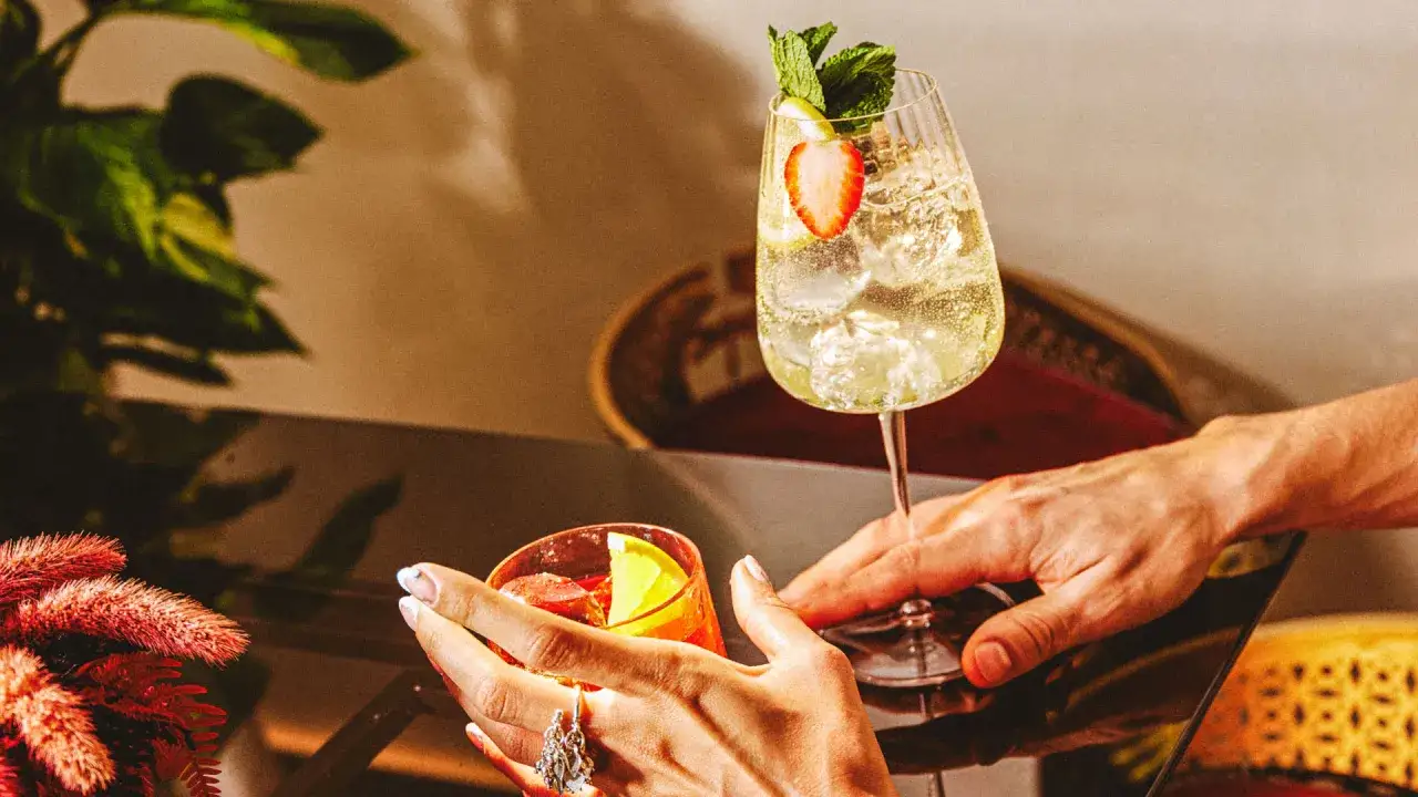 Deux mains tendent des cocktails rafraîchissants, un verre de martini bitter rouge et un autre pétillant garni de fraises et de menthe.