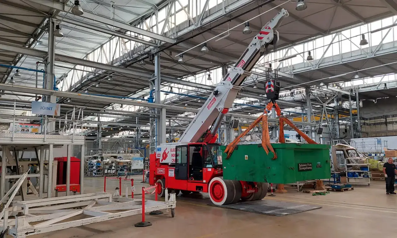 W hali produkcyjnej trwa przemieszczanie maszyn. Czerwony dźwig elektryczny podnosi zieloną skrzynię.