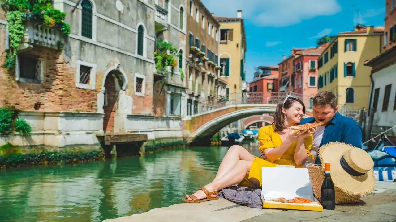 Un couple partage une pizza au bord d'un canal &agrave; Venise, ville des amoureux. Les b&acirc;timents color&eacute;s et un pont cr&eacute;ent un d&eacute;cor romantique.