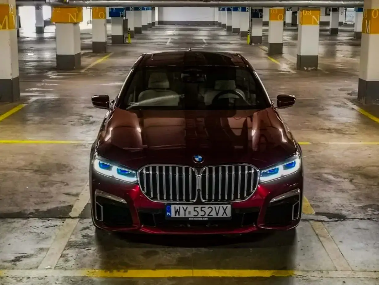 Lśniące, bordowe BMW 750i z niebieskimi światłami dziennymi stoi na parkingu podziemnym.