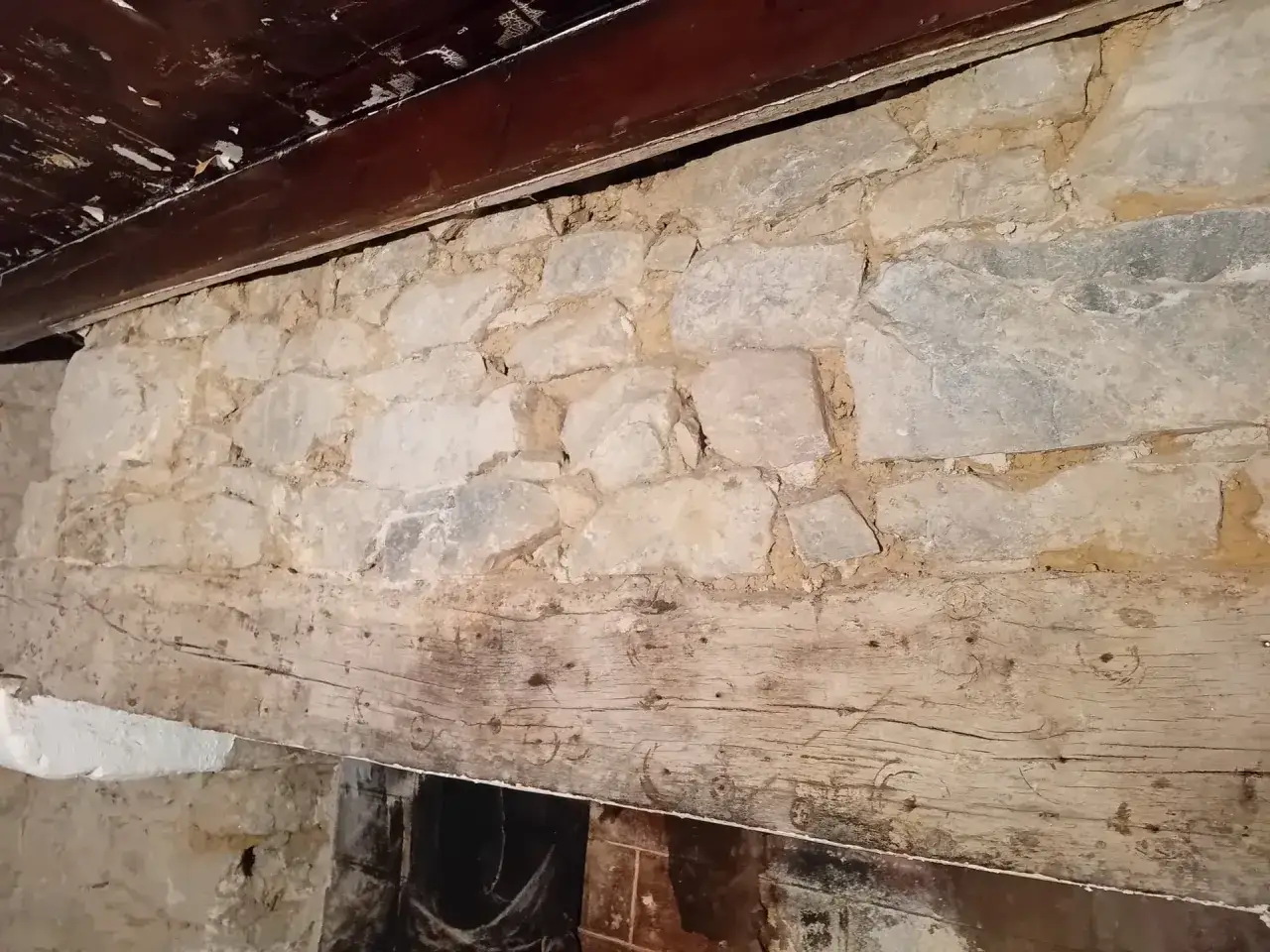 Gros poutre en bois patinée sous un mur en pierre, vestige d'une ancienne bâtisse.