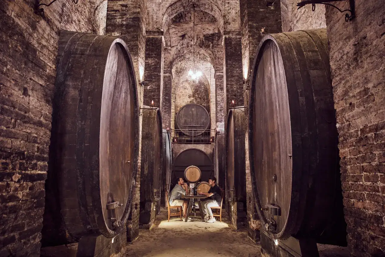 Due uomini seduti a un tavolo tra botti giganti nelle cantine storiche di Montepulciano, un'atmosfera suggestiva.