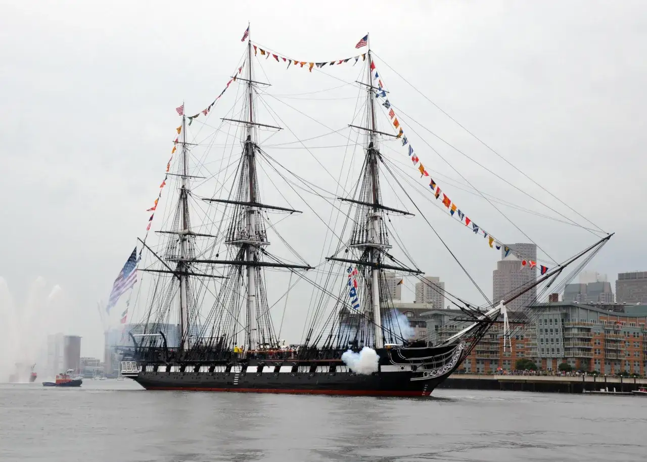 Le majestueux shtandart bateau, orné de drapeaux colorés, navigue fièrement sur l'eau, salué par des canons fumants.