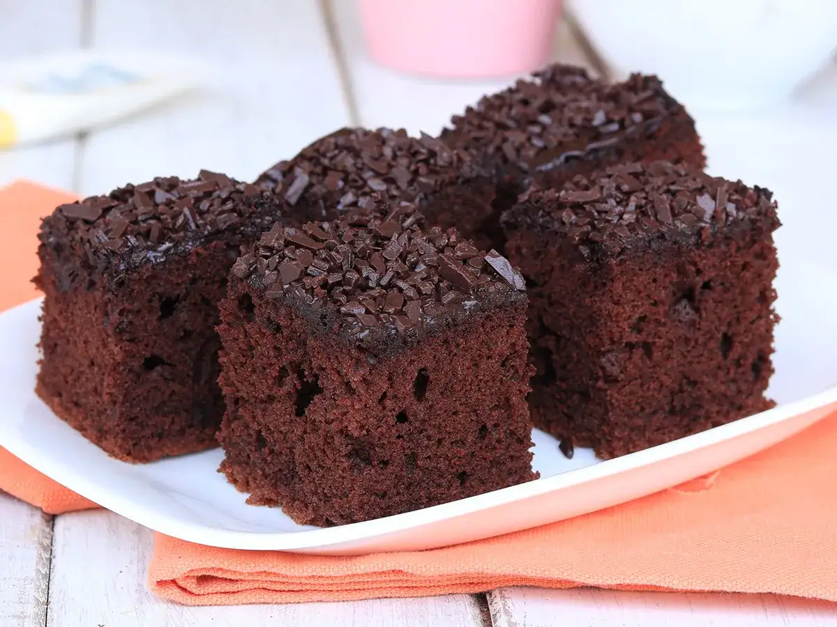 Quattro fette di torta al cioccolato morbida e umida, ricoperte di granella di cioccolato, su un piatto bianco con tovagliolo arancione.