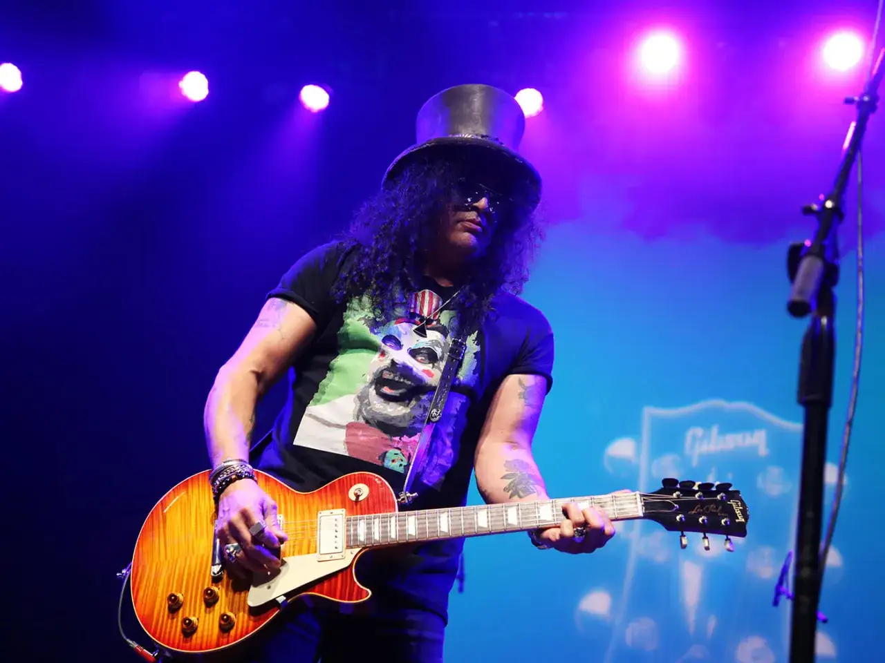 Le guitariste Slash, coiffé d'un chapeau haut-de-forme, joue sa Gibson sur scène sous les lumières bleues.
