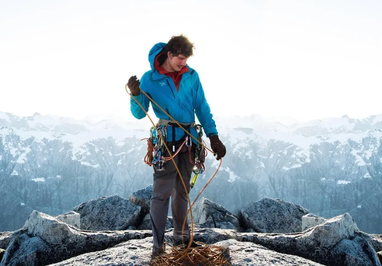 Grimpeur en veste shell, c'est quoi ? Il manipule une corde sur un sommet rocheux, avec des montagnes enneigées en arrière-plan.