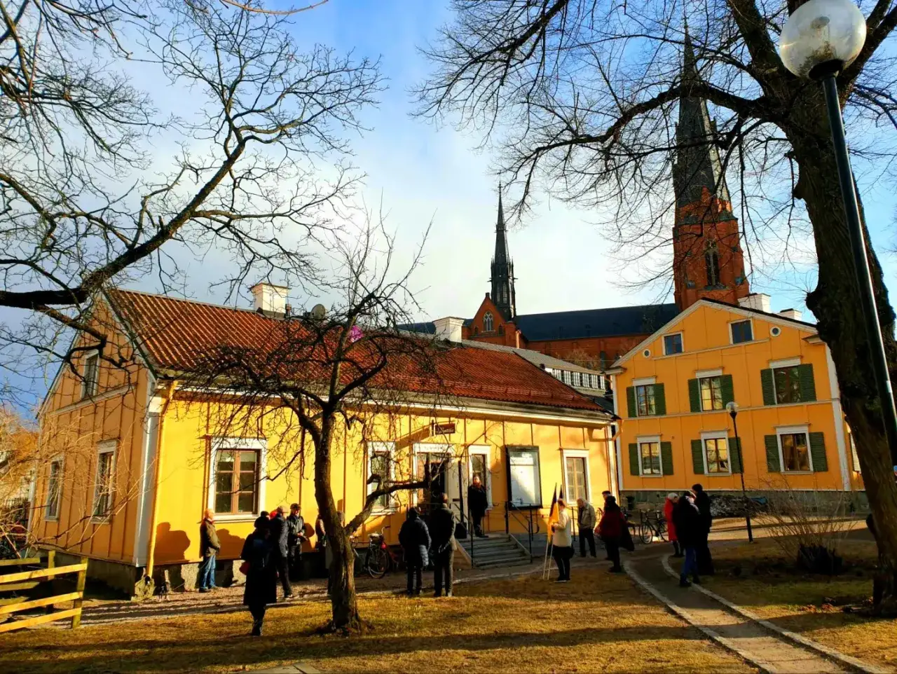 Gula hus och Uppsala domkyrka i bakgrunden. En grupp m&auml;nniskor samlas utanf&ouml;r, kanske f&ouml;r ett bes&ouml;k p&aring; litteraturens hus Uppsala.
